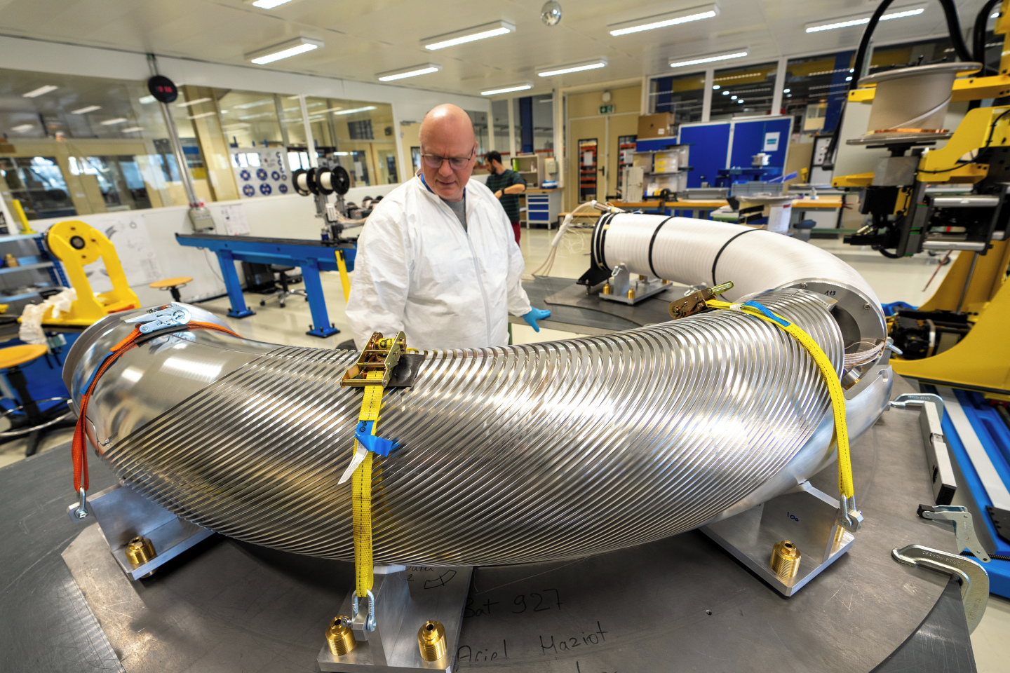 In a physics lab, a person in a protective suit observes the curved outer former which is secured with straps on a metal platform. The shape and grooves in the surface vaguely resemble a pasta shape. The inner former is wrapped in white and ready to be inserted