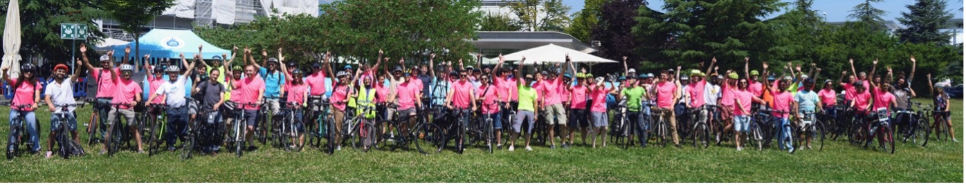 A large number of cyclists stand in a row on or next to their bicycles, many in pink t-shirts and with their arm/arms in the air.
