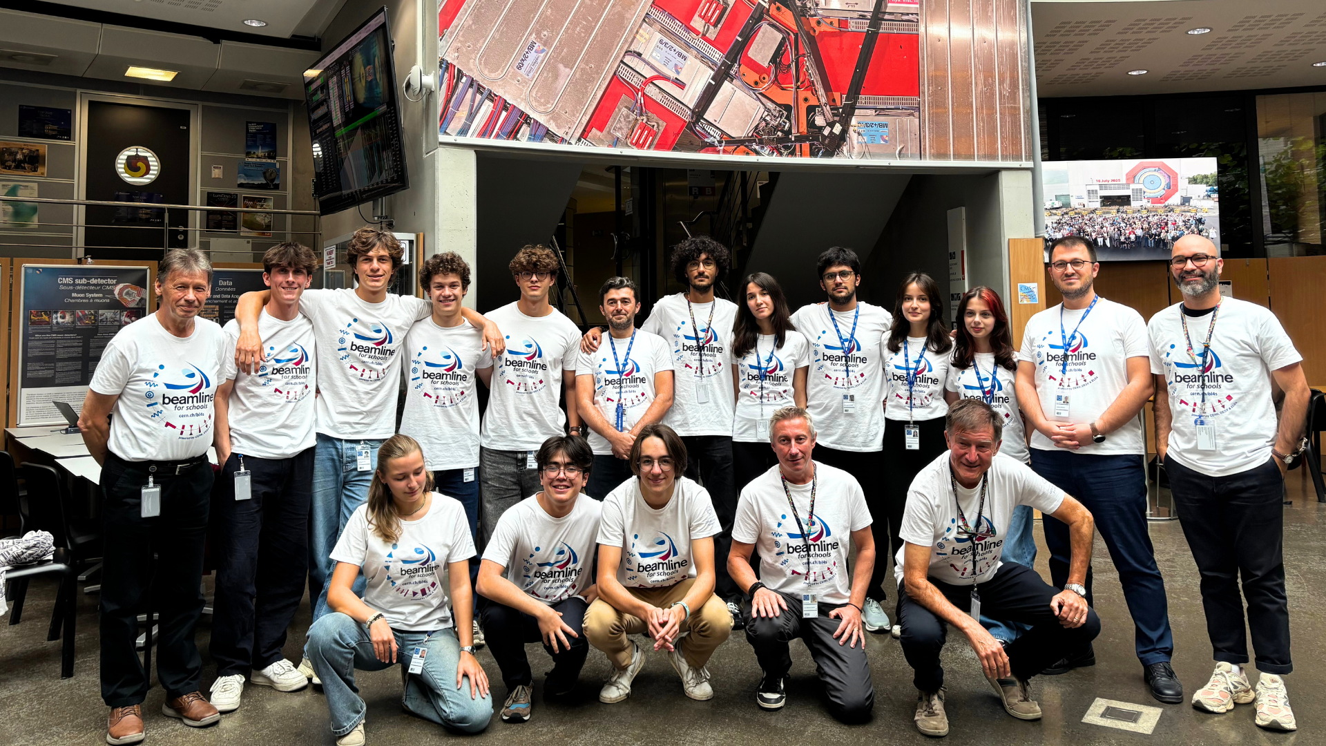 A group of people in two rows. The first row is kneeling and the back row is standing. They are smiling and friendly with each other. They are all wearing white branded t-shirts.