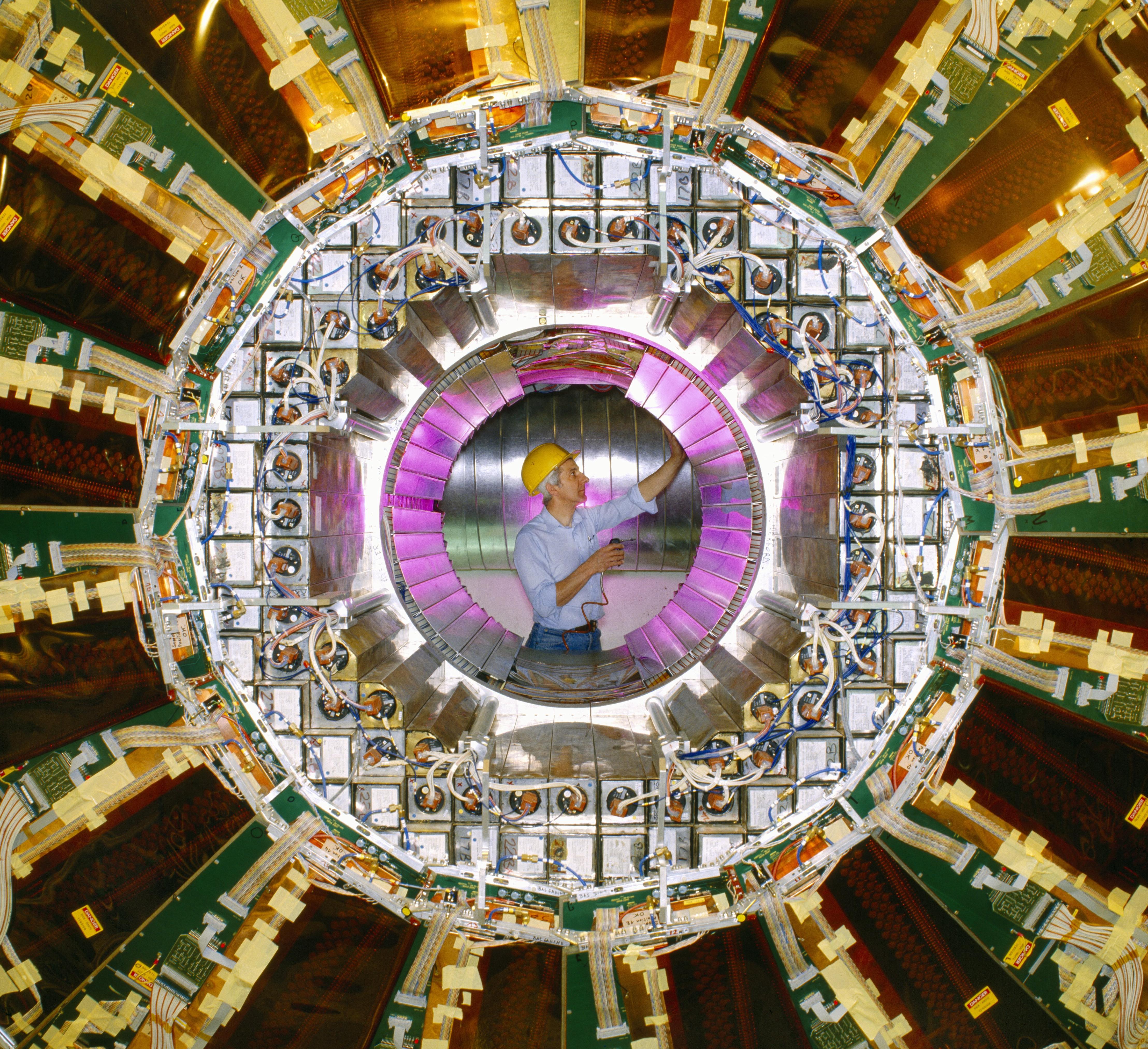 The OPAL detector is a large cylinder with layers of metal in gold and green with wires connecting it the magnets together. The centre of the cylinder is hollow and at the end we can see a man in a blue shirt and yellow hat working on the detector.