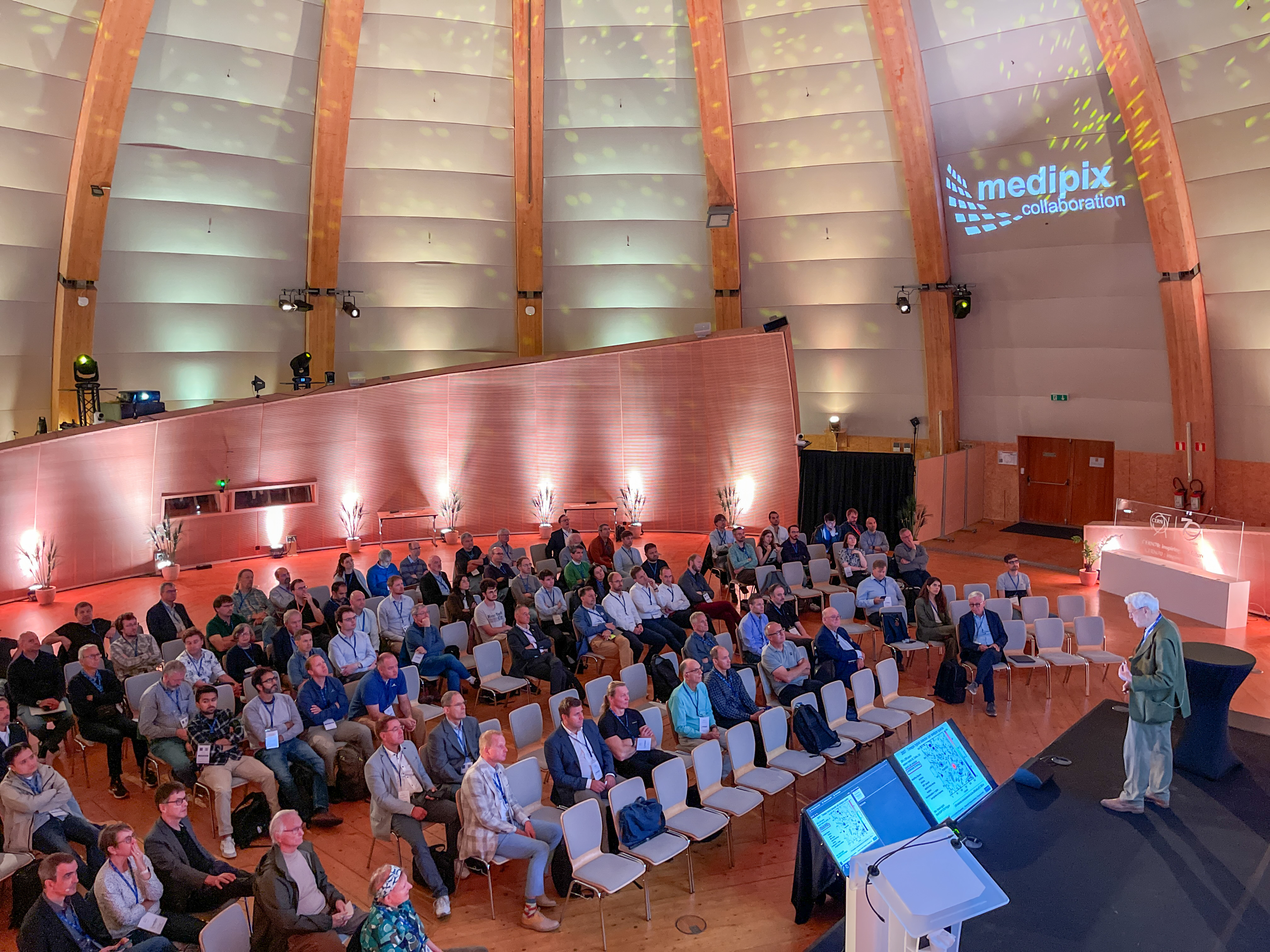 A crowd of people sat in a large dome-shaped building are watching a talk by a man on a stage in the lower right-hand corner.