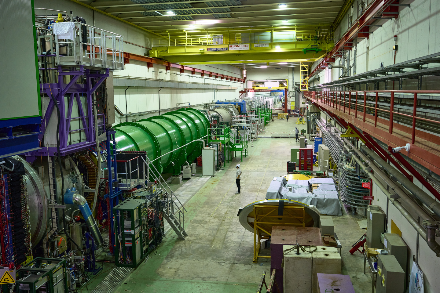 A view down a large cavern. On the left hand side, there is a large green metal tube housing part of the NA62 experiment. A man in a hard hat is standing next to it for scale. He is about a quarter the height of the tube.