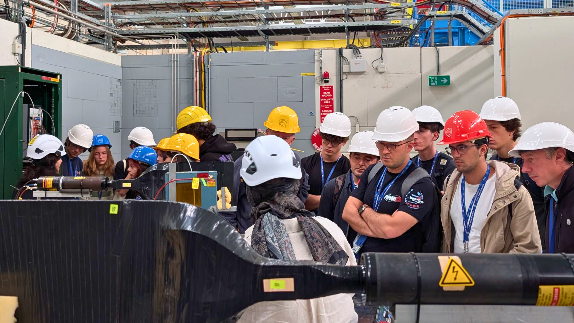 A crowd of people are in a large warehouse, all wearing hard hats. They are crowded around to look at what a guide is showing them.