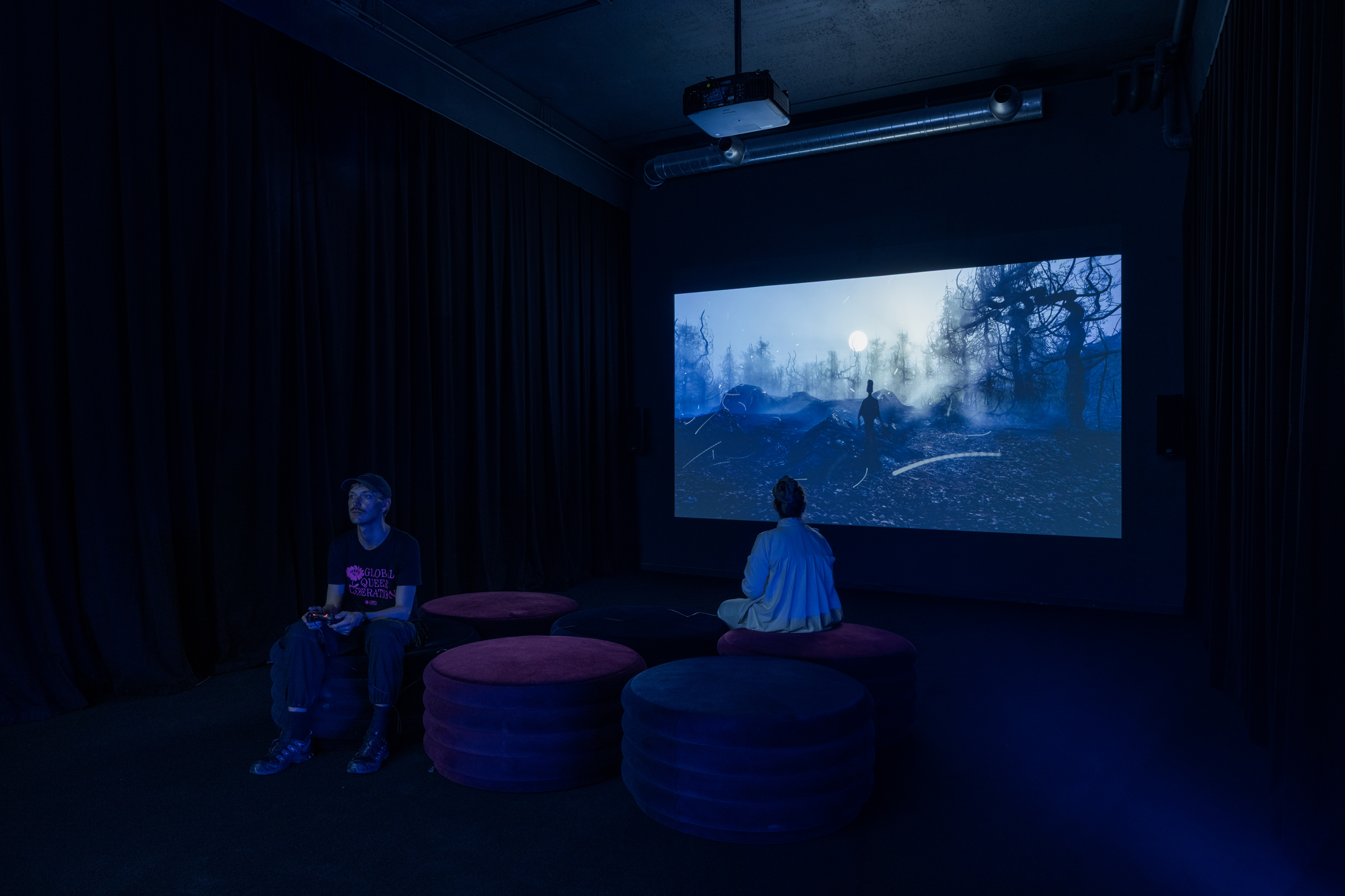 Two people sitting down with a game controller in their hand, facing opposite directions. In the back, you see on a screen the game they are playing