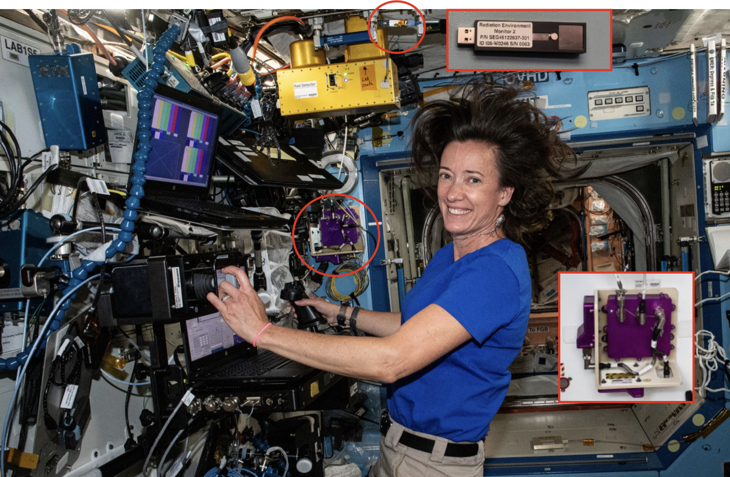 A woman in a blue shirt is smiling towards the camera. She is working at a complex-looking station with two laptops with many tubes and cables. Her hair is floating around her due to the zero gravity.