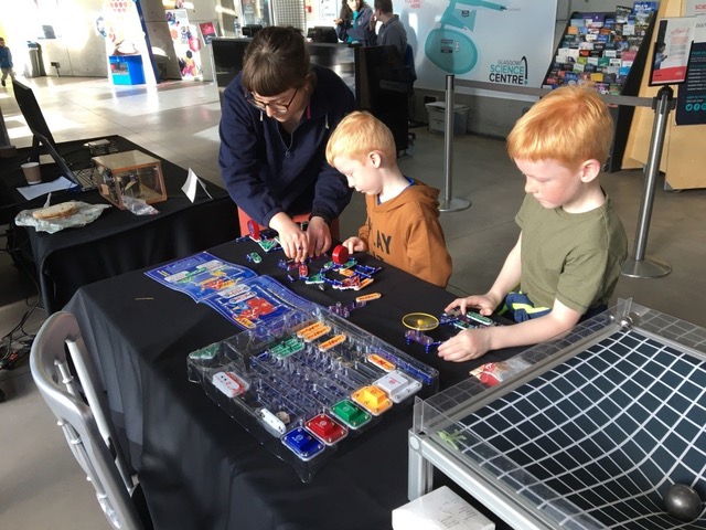 Two children are looking intently at interactive exhibitions while an adult guide is explaining something to them.