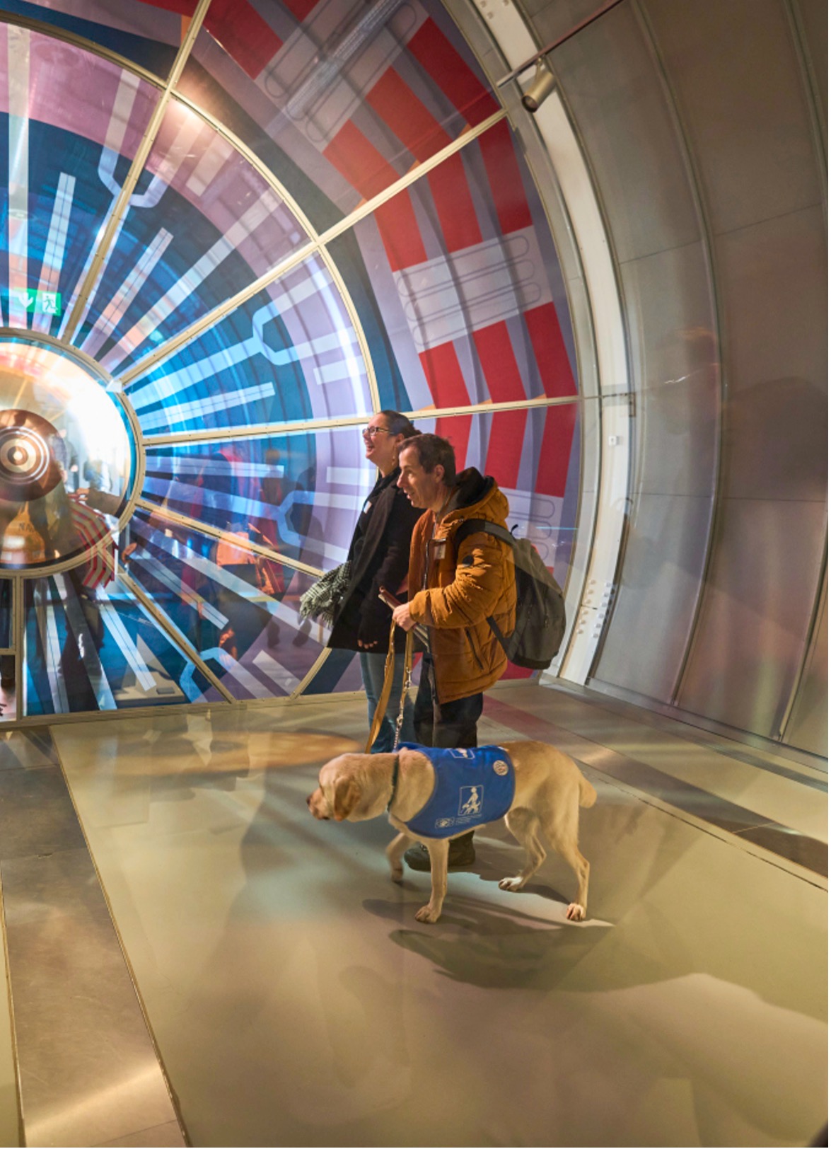 A visitor from ABA touring the “Discover CERN: Collide” exhibition with his guide and guide dog. In the background is a transluscent circular window with red and blue sections to show a slice of a CERN detector