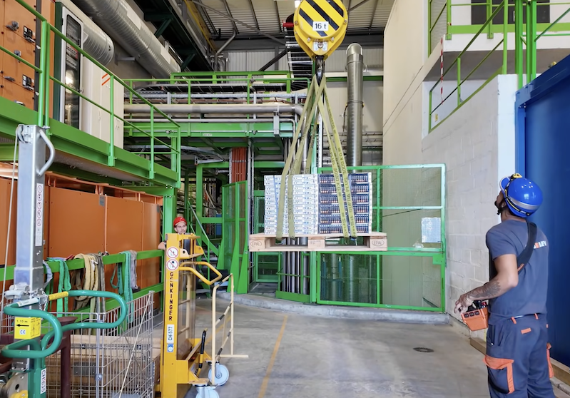One worker in a hard hat controls a crane as it lowers a pallet with computing equipment. Another worker, also in a hard hat, watches on.