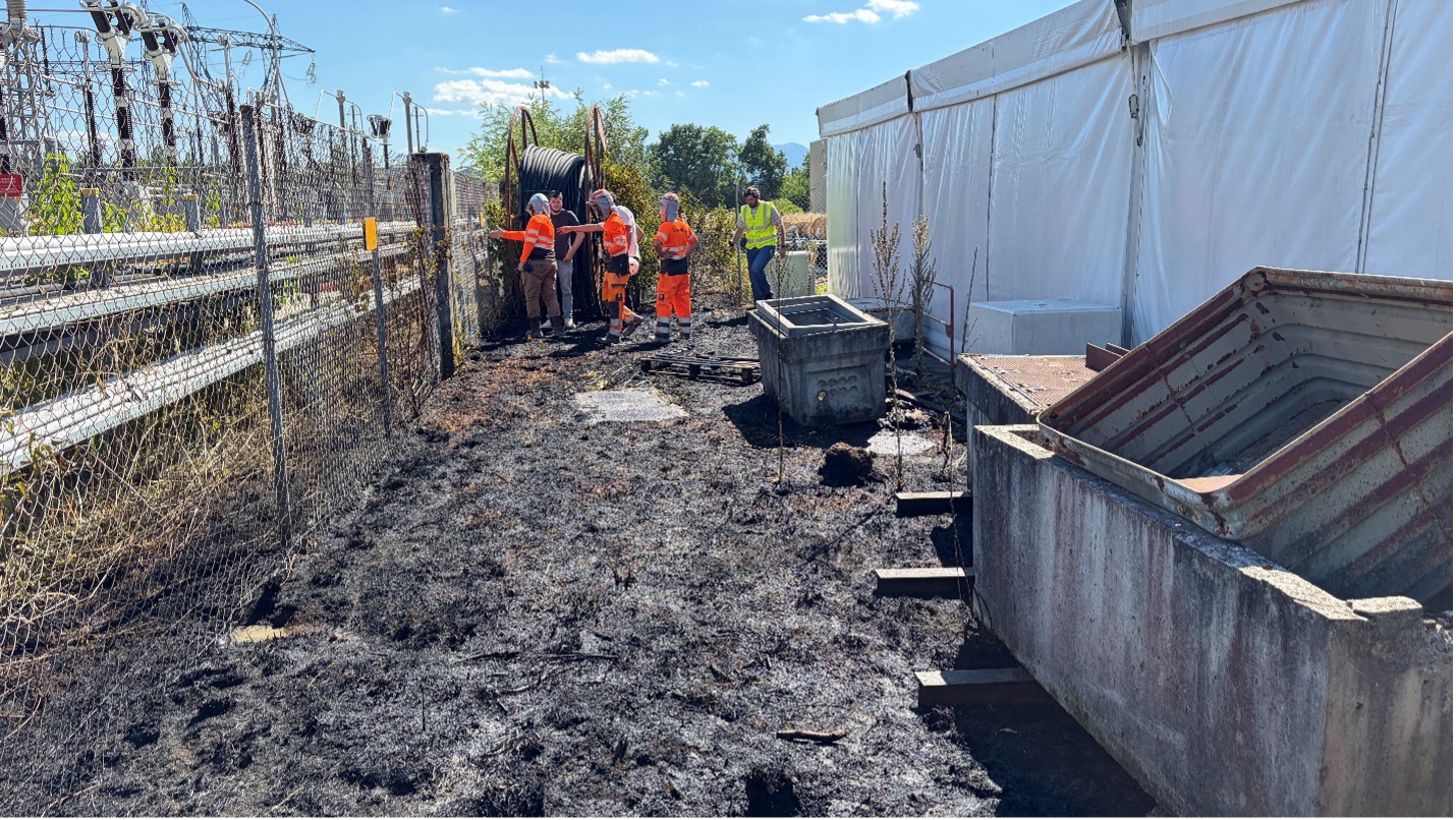 A small patch of burnt vegetation, with members of the CERN fire service in orange high vis jackets and hoods, accompanied by other CERN staff.