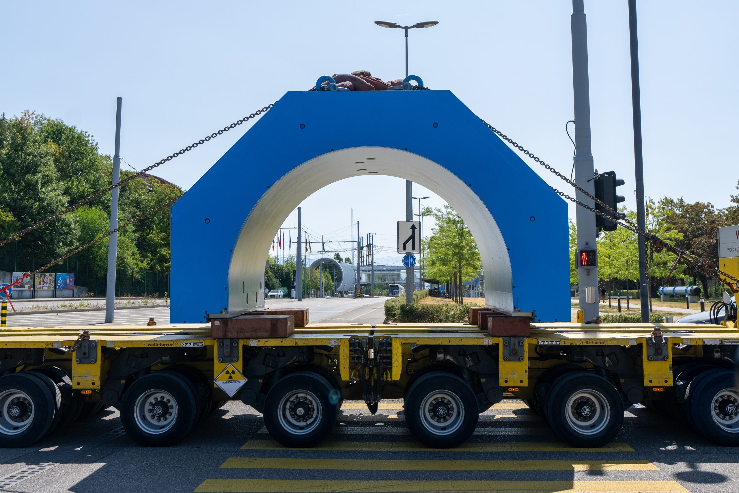 A very large blue arch-shaped magnet travelling on the back of an open lorry. Through the magnet you can see a glimpse of CERN science gateway, and to the left of the magnet the Globe of Science and Innovation, mostly obscured by trees