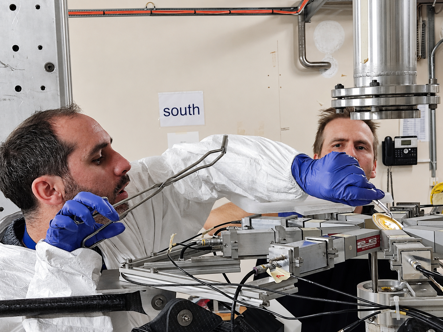Two men, one in blue gloves handling long tongs, work on piece of equipment made of metal sections and wires
