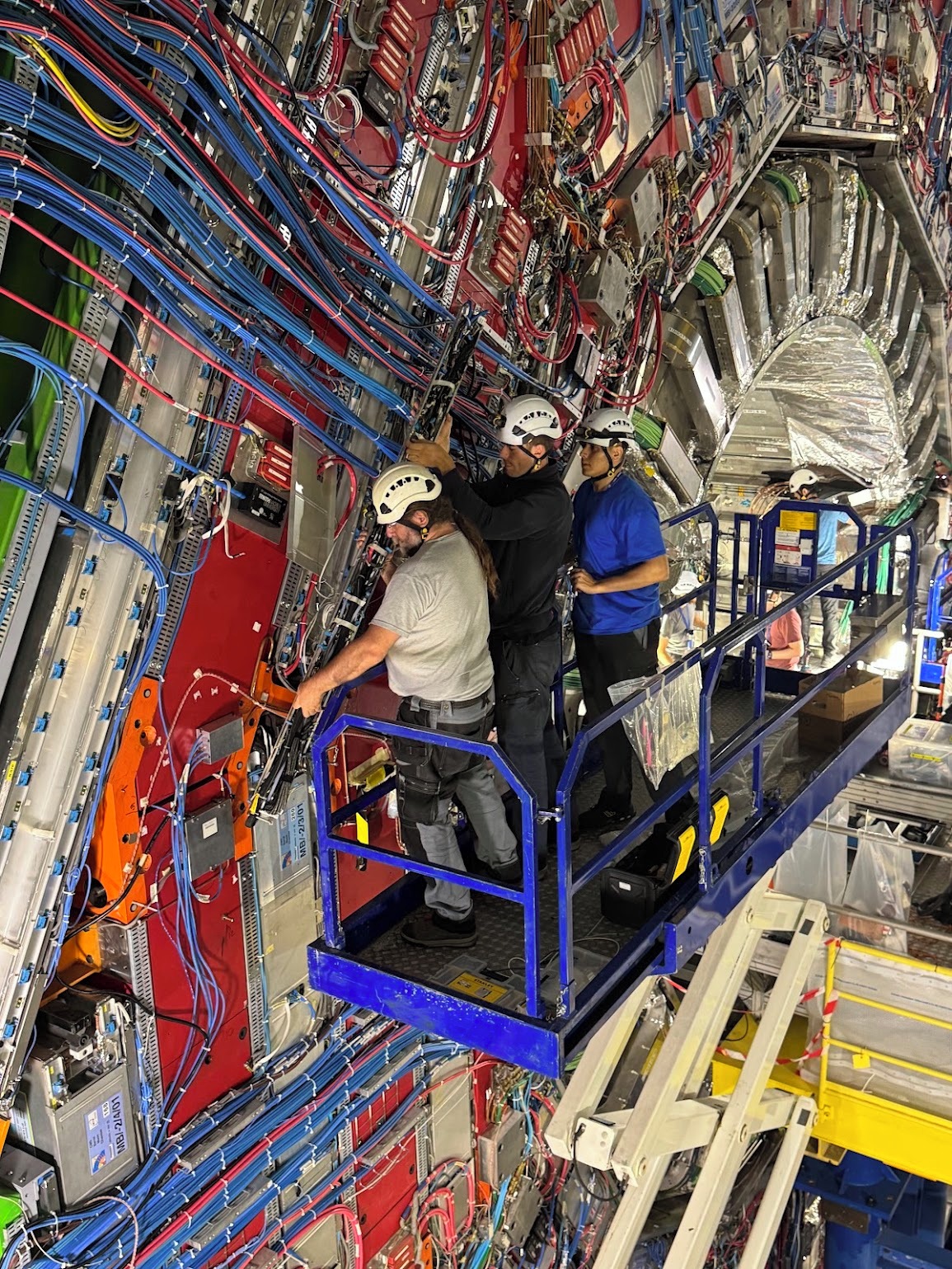 Three men on a blue crane are installing some equipment on one of the giant red slide of the CMS detector